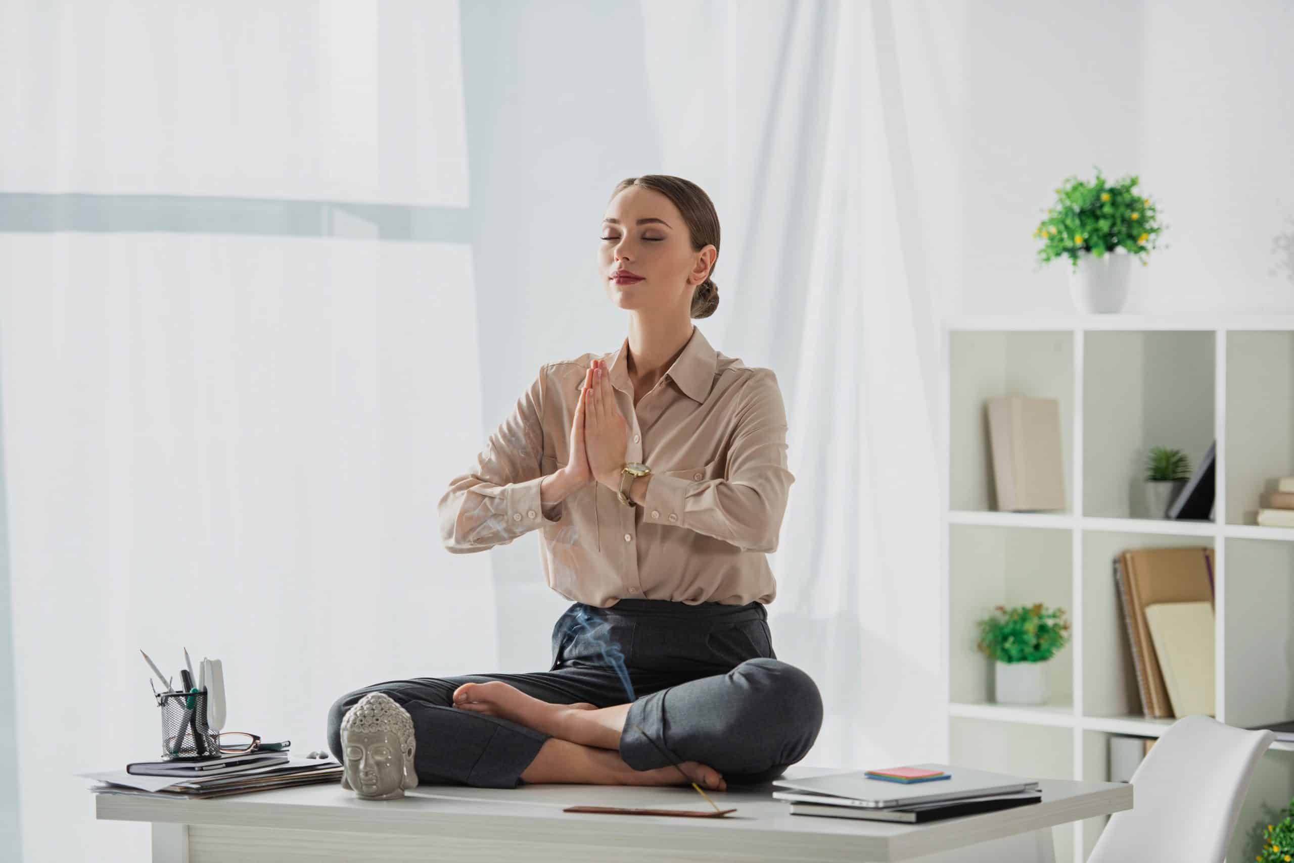 visuel d'une femme assise sur son bureau en position du lotus et faisant le geste namasté avec une tête de buddha et un baton d'encens posés sur le bureau