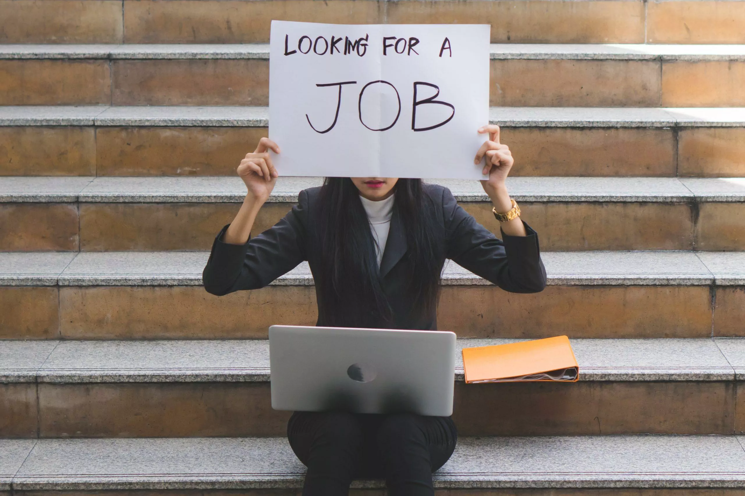 visuel d'une femme assise sur des marches avec un ordinateur portable sur les genoux et tenant une pancarte devant son visage où il est écrit looking for a job