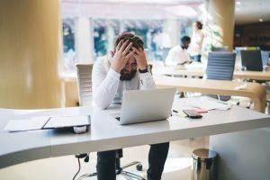 un homme se tenant la tête entre les mains, assis à son bureau devant son ordinteur