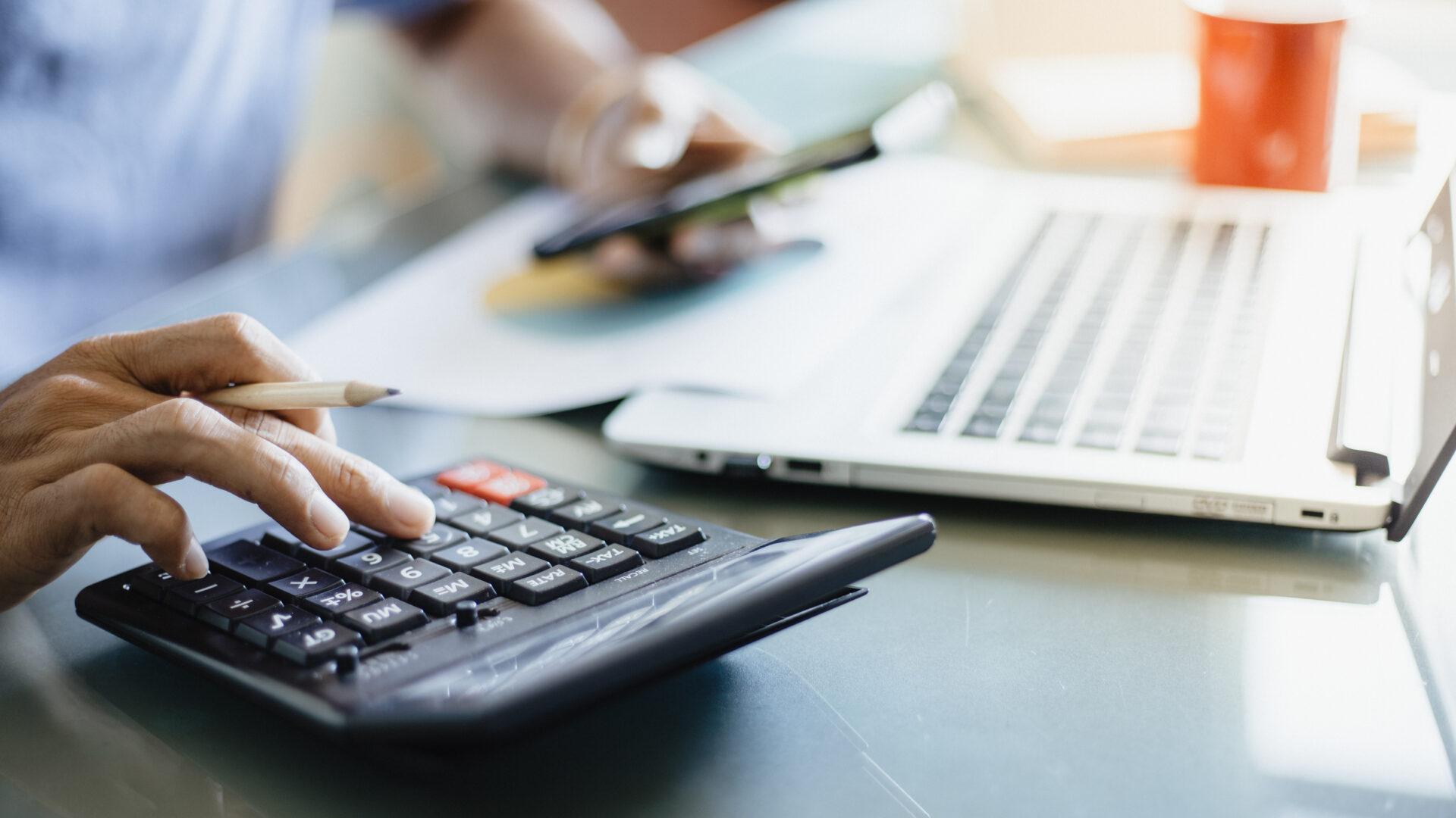 visuel d'un homme utilisant une calculatrice de la main droite et son téléphone de la main gauche devant un ordinateur portable